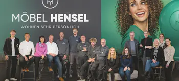 Gruppe von Personen steht und sitzt vor Wand mit Schriftzug Möbel Hensel und Wohnen sehr persönlich sowie großem Portrait rechts
